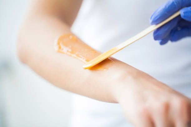 Person applying warm wax on an arm using a wooden spatula during a waxing procedure.